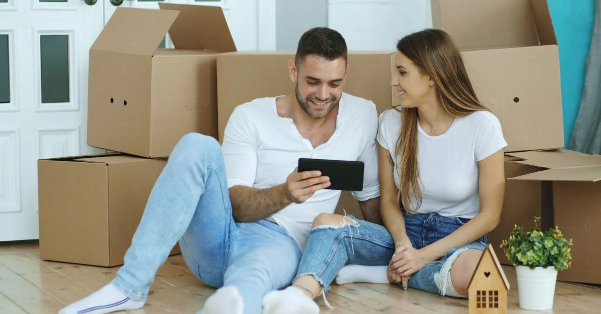 Jeune couple déballant joyeusement dans leur nouvelle maison, consultant quelque chose sur une tablette.