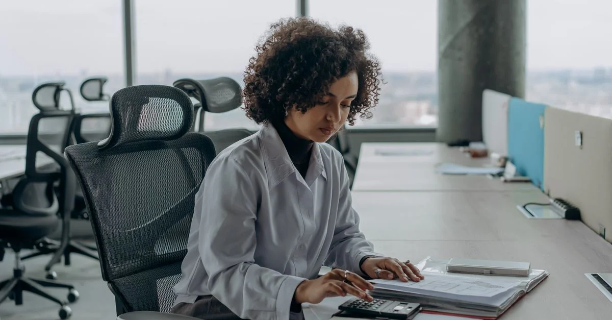 Une femme professionnelle avec des cheveux bouclés calculant des documents dans un cadre de bureau moderne.